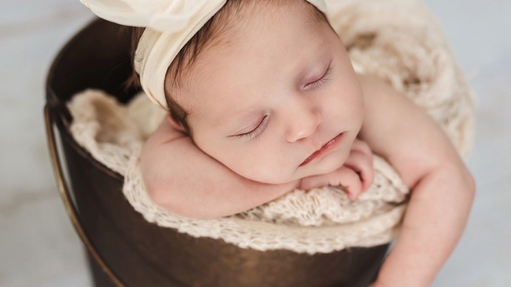 Newborn sleeping in a metal bucket, cream cloth headband and white knit lining around her, resting her cheek on her arms. Styled posed portrait.