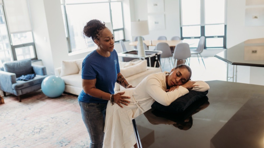 A doula in a navy tee presses on the low back of a laboring woman in a white robe who is bent over a pillow on the kitchen counter. Wide view of the bright high-rise apartment.
