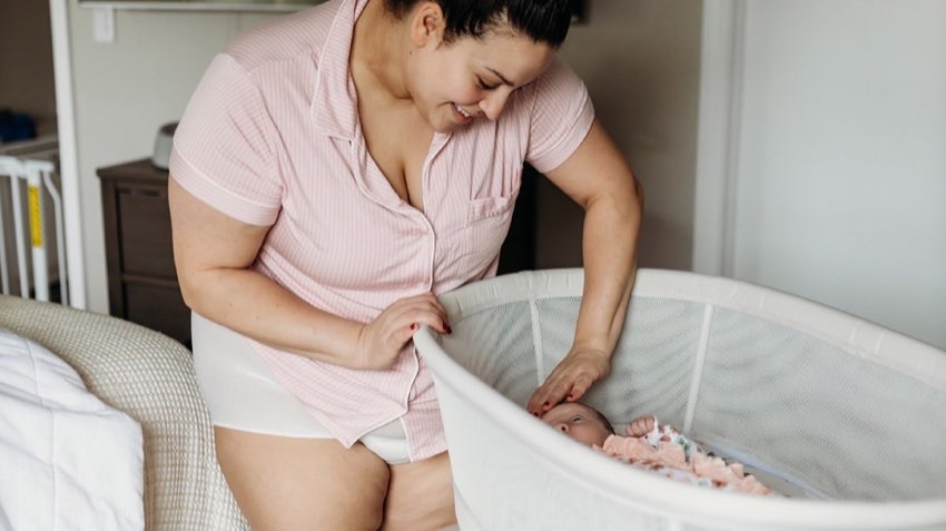 Mom laughs warmly as she lays her newborn down in the bedside bassinet. Joyful, candid postpartum scene.