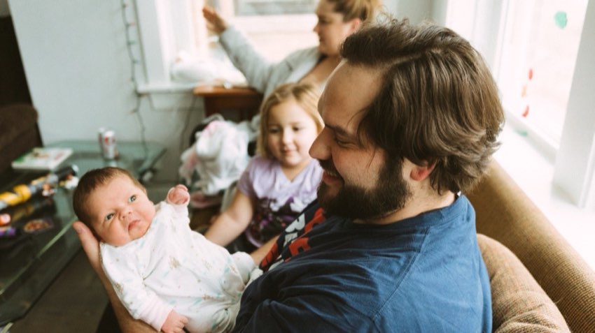 Dad smiling broadly while cradling his newborn daughter (peach skin, wide eyes); older daughter looks on, mom in robe blurred behind. Candid family moment.