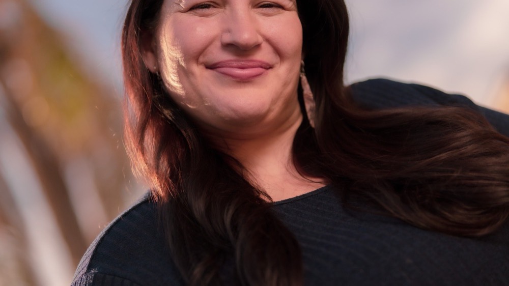 Outdoor portrait of Anna in a navy ribbed top with a soft closed-mouth smile, hair draped over one shoulder. Bright blue sky and palms blurred behind her.